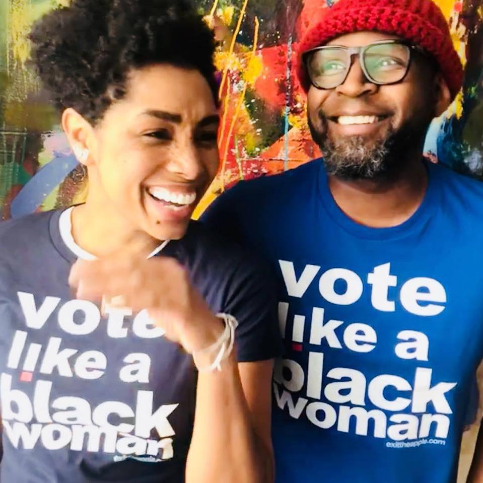 image of two smiling black people, a woman with short hair and a man in a red hat, looking in different directions. text on their shirts reads 'vote like a black woman'